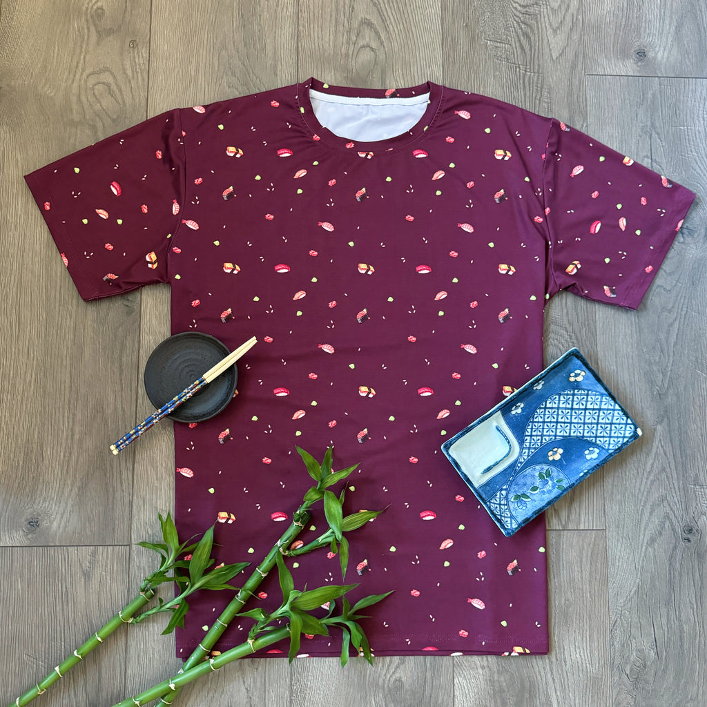 Maroon T-shirt with an all-over sushi pattern, displayed on a wooden floor with accessories like a book, bamboo plant, and chopsticks for a styled look.