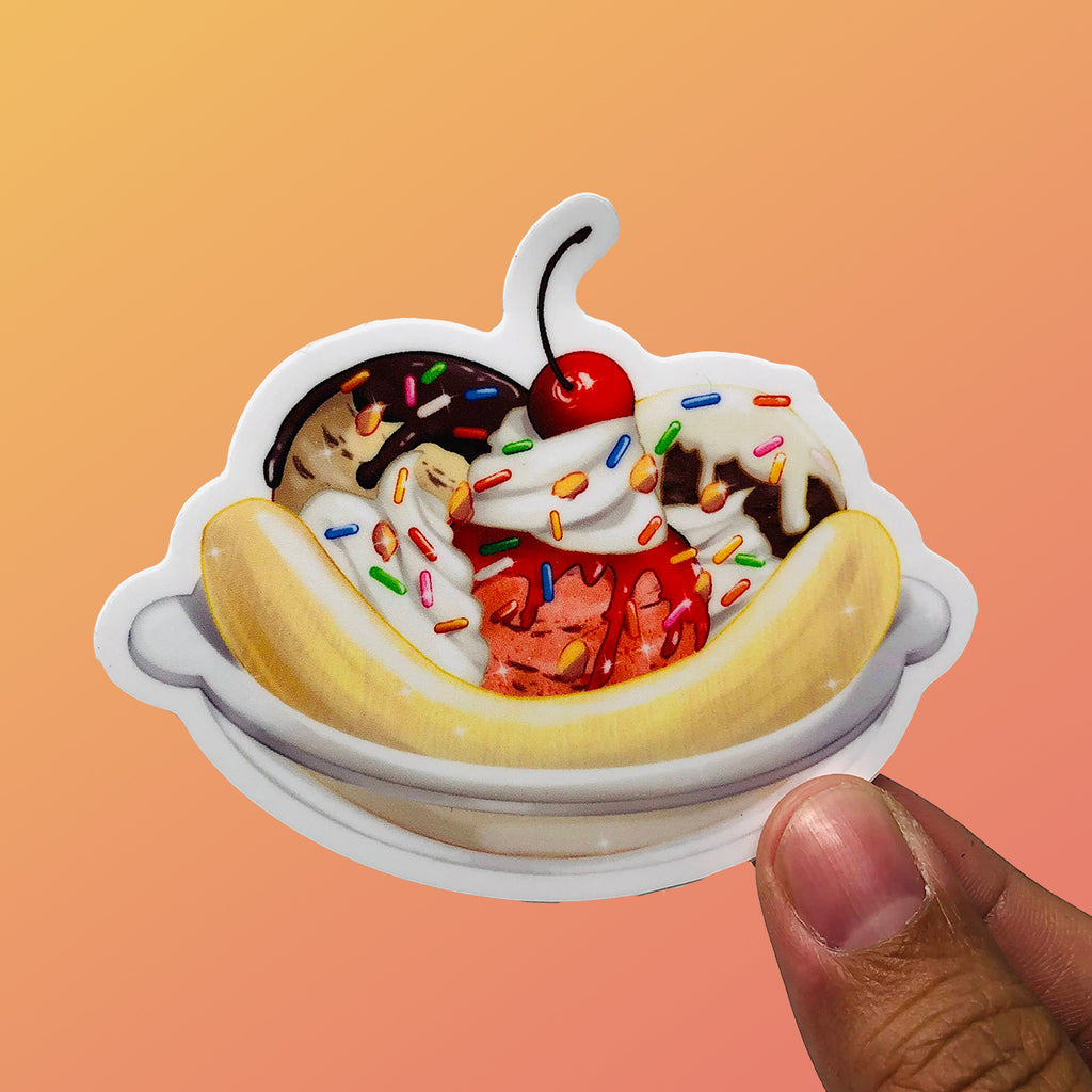 Colorful vinyl sticker featuring a detailed banana sundae with ice cream scoops, sprinkles, and a cherry on top, held against a gradient orange background